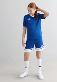 Maillot de sport bleu avec un col en V blanc, assorti à un short avec des rayures blanches et bleues. Chaussettes blanches et crampons.