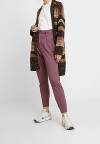 Pletený kardigan s hnědými a krémovými pruhy, bílá košile na knoflíky, přiléhavé malinové kalhoty a sportovní tenisky s bledými akcenty.