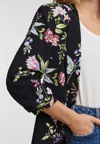 Blazer negro de manga larga con flores en rosa, azul y blanco, llevado sobre una camiseta blanca, combinado con vaqueros azules de mezclilla.