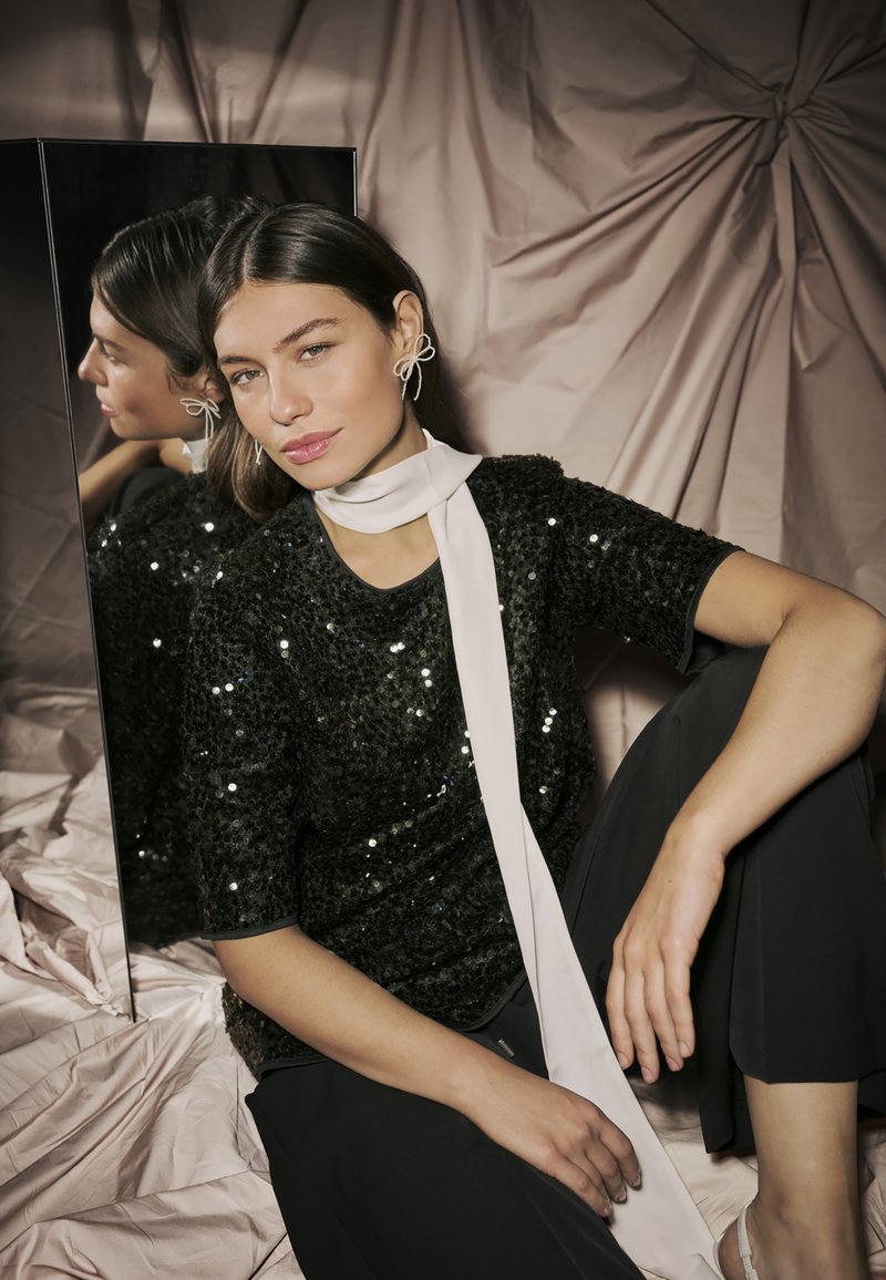 Melnas sekvīnu blūze ar īsām piedurknēm, kopā ar gaišu satīna šalli. Modelis pozē sēdošā pozīcijā blakus atstarojošai virsmai uz mīkstas drapētas auduma.