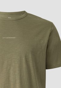 T-shirt vert olive à col rond avec manches courtes et petit texte beige "LET'S SYNERGIZE !" sur le côté gauche de la poitrine.
