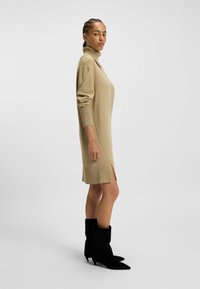 Beiges gestricktes Kleid mit hohem Kragen, gerippten Bündchen und seitlichen Schlitzen. Kombiniert mit schwarzen spitzen Ankle Boots.