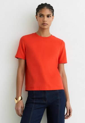 Mlada ženska s pletenimi lasmi nosi enostavno svetlo oranžno kratko srajco, temne kavbojke iz jeansa in zlato skulpturalno zapestnico, stoji ob belem zidu.