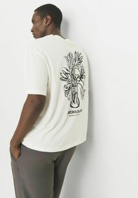 T-shirt en coton blanc avec une illustration de vase floral noir au dos. Il possède des manches courtes et une coupe décontractée. Logo accent sur le bas.