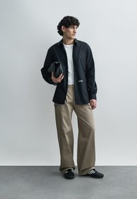 Črna srajca s potiskanimi gumbi čez belo majico, bež široke hlače, v rokah drži črno torbo. Črni čevlji z belimi črtami.