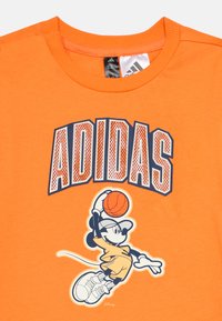 Πορτοκαλί βαμβακερό μπλουζάκι με την επιγραφή "ADIDAS" σε έντονα γράμματα. Σχέδιο με ένα καρτούν χαρακτήρα που ντριμπλάρει μια μπάλα του μπάσκετ κάτω από το κείμενο.