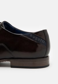 Chaussure habillée en cuir marron avec une finition brillante, comportant un accent en tissu bleu contrasté avec le logo "bugatti" sur le côté. Semelle en bois.