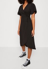Černé zavinovací šaty s krátkými volánkovými rukávy, stahovaným pasem a asymetrickým lemem, v kombinaci s černými vysokými teniskami.