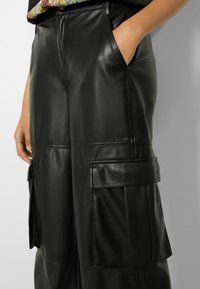 Osoba nosząca czarne, sztuczne skórzane spodnie cargo z bocznymi kieszeniami, z rękami częściowo schowanymi w kieszeniach w pasie.