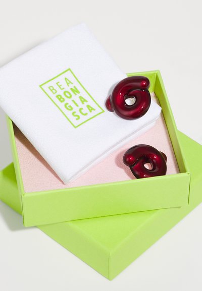 Κόκκινες ρητίνες σκουλαρίκια σε σπειροειδή σχέδιο, εκτεθειμένα σε ένα πράσινο κουτί με ροζ εσωτερικό και λευκ�ή υφασμάτινη επένδυση με το εμπορικό σήμα.