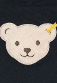 Navyblaues Hemd mit einem plüschigen, hellfarbenen Bärenkopfabzeichen, das braune gestickte Details und ein kleines, gelbes Akzentetikett aufweist.