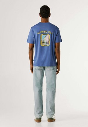 Hombre con camiseta azul con estampado de Notting Hill, jeans azul claro y zapatillas beige, de pie de espaldas sobre fondo blanco.