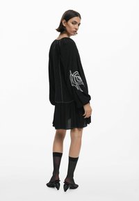 Das schwarze Langarmkleid verfügt über ein besticktes Blumenmuster, einen gestuften Rock und kontrastierende weiße Nähte. Kombiniert mit transparenten schwarzen Socken und Absatzschuhen.