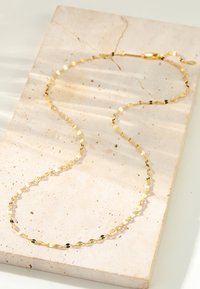 Złoty naszyjnik o geometrycznym wzorze, z naprzemiennymi płaskimi i zaokrąglonymi ogniwami. Gładka tekstura, z zapięciem na jednym końcu, położony na jasnej kamiennej powierzchni.