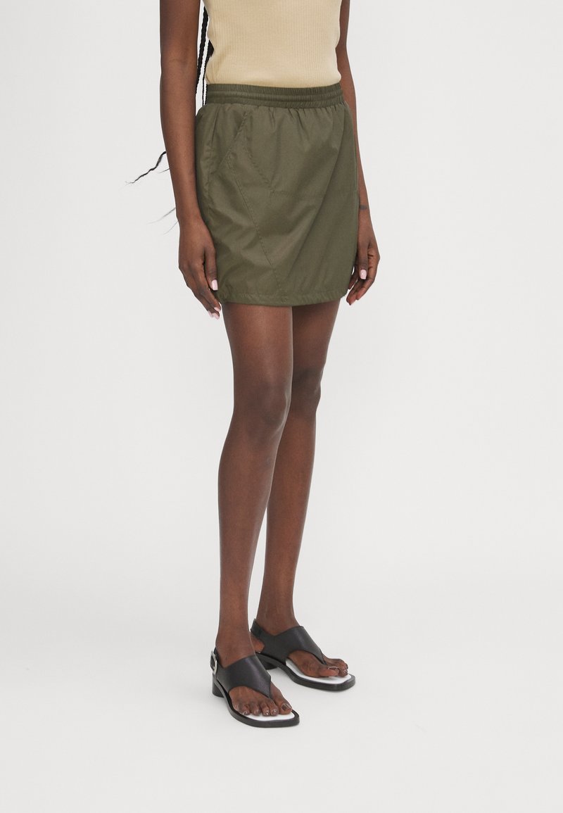 Olive green, lightweight, elastic-waist skirt with side pockets, paired with black sandals featuring a sleek design and silver accent.