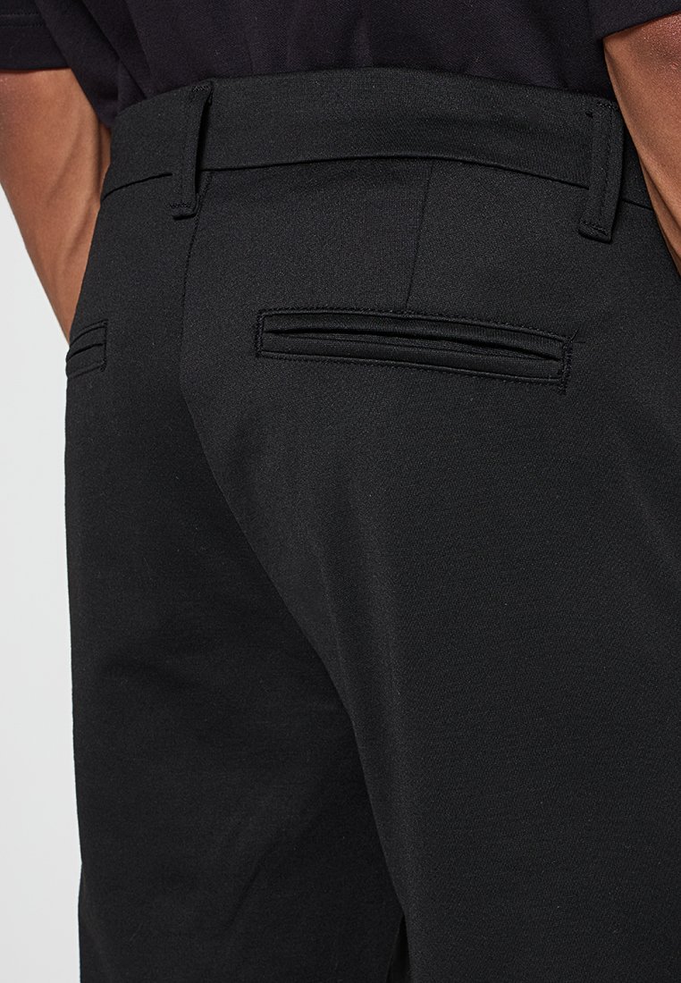 Schwarze, maßgeschneiderte Hose aus glattem Stoff mit schlanker Passform und zwei Taschen auf der Rückseite. Das Design betont einen sauberen, professionellen Look.