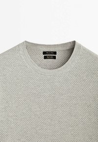 Pull en maille texturée gris clair à col rond avec col côtelé et étiquettes de marque noires à l'intérieur du col.