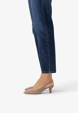 Sötétkék farmernadrágot viselő lábak, bézs színű, velúr slingback kisméretű sarokcipőkkel, fehér háttéren állva.