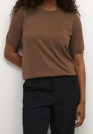 Kvindelig med krøllet hår iført en kortærmet brun striktrøje og sorte bukser, står op ad en hvid væg med den ene arm bag ryggen.