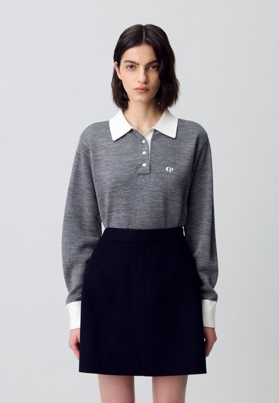 Polo de punto gris con cuello blanco y cuatro botones, combinado con una falda negra de talle alto. Presenta un sutil logo en el pecho.