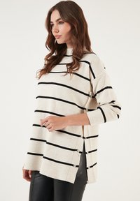 Maglione a righe color crema con strisce orizzontali nere, dotato di un collo alto, bottoni laterali e una vestibilità oversize.