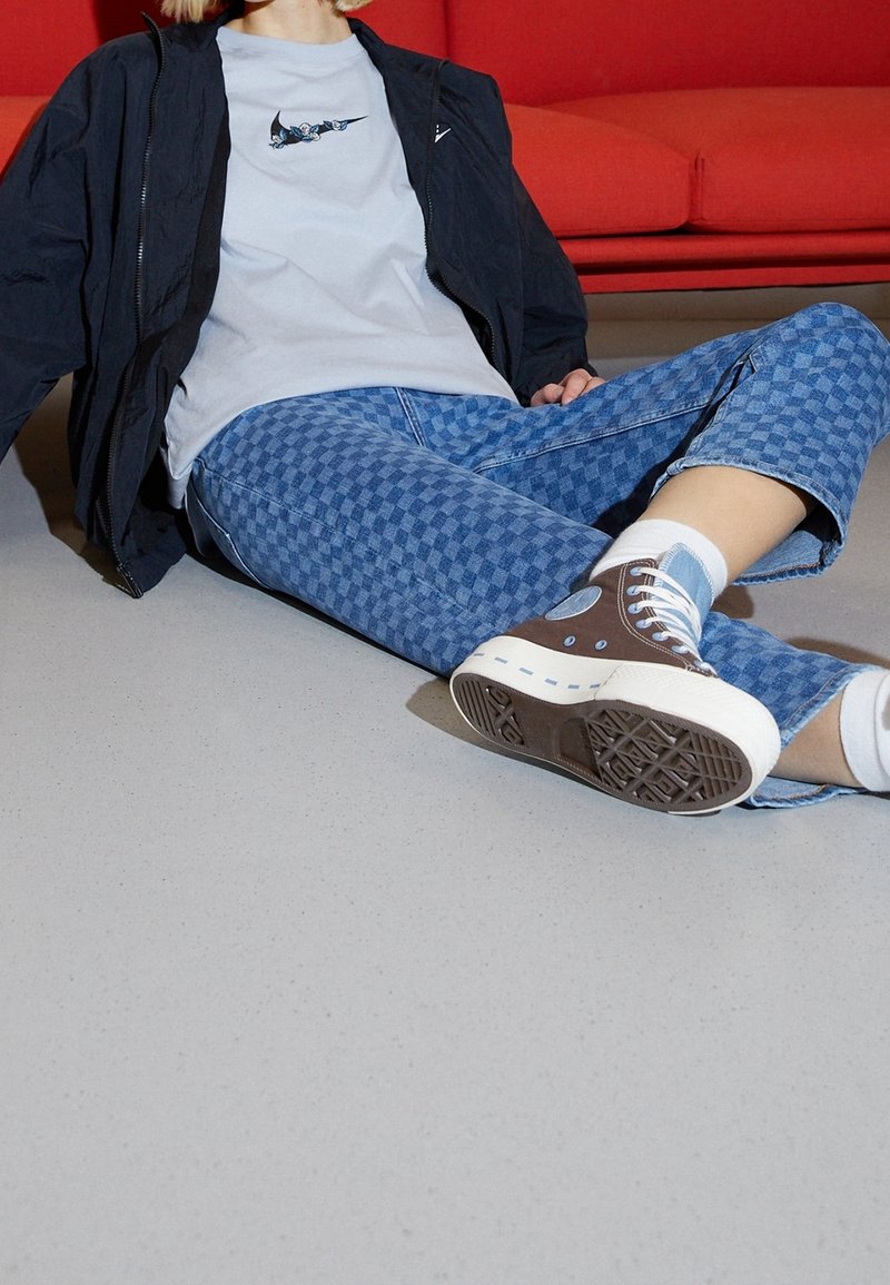 Bruna och blå hög-top sneakers med vit sula; blå rutiga jeans; ljusgrå t-shirt; svart jacka; vita strumpor; slät grå golv.