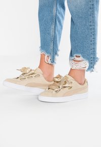Baskets en daim beige avec lacets en satin, semelle en caoutchouc blanche et détail du logo doré. Portées avec un jean en denim bleu déchiré.