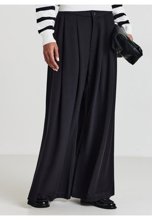 Persona che indossa pantaloni larghi neri, scarpe nere, un maglione bianco con strisce nere, mentre tiene una pochette nera.