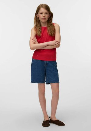 Young person with long hair wearing a red sleeveless top, blue denim shorts, and dark slip-on shoes standing with arms crossed.