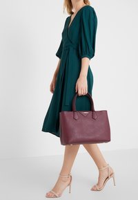 Burgunderfarvet lædertaske med struktureret form, to håndtag og sølvlogo. Kombineret med en blågrøn kjole og beige sko med stropper.