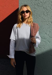 Szara, oversize'owa koszulka warstwowa na długą białą koszulę, zestawiona z czarnymi legginsami. Modelka trzyma w ręku brązowo-pomarańczowy szalik w kratę.