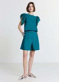 Haut teal avec des manches en plumes, associé à un short plissé teal assorti. Le mannequin porte des chaussures à talons argentées. Fond uni.