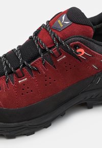 Chaussure de randonnée en daim rouge et mesh noir avec une semelle en caoutchouc robuste, dotée de trous de ventilation bien visibles et de lacets à motif.