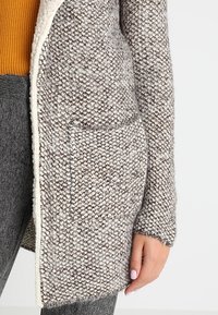 Pitkä neulecardigan vaaleanruskeissa ja harmaissa sävyissä, jossa on tekstuurinen kangas, avoin etuosa ja sivutasku kontrastivärillä.