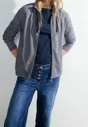 Frau trägt ein marineblaues T-Shirt, eine graue Kapuzenjacke mit Reißverschluss und Kordelzug sowie hoch taillierte blaue Jeans mit vier Knöpfen.