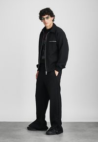 Homme aux cheveux bouclés portant une veste zippée noire, un pantalon large assorti, des lunettes de soleil et des baskets noires, se tenant devant un fond uni.