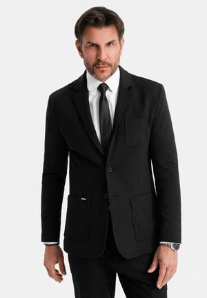 Mann mit kurzem dunklen Haar und Bart, der eine schwarze Sakkojacke, ein weißes Hemd, eine schwarze Krawatte und eine Armbanduhr trägt und vor einem schlichten weißen Hintergrund steht.