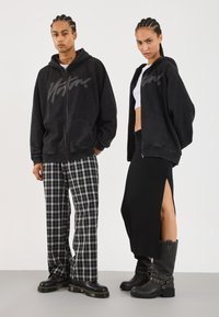 Black zip-up hoodie with logo, paired with checkered pants. Model on right wears a black skirt with a slit and buckled boots.