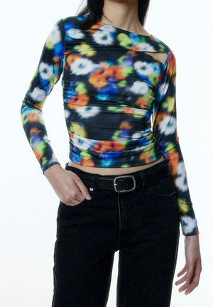 Kobieta ubrana w dopasowaną, długorękawową, wielokolorową bluzkę w kwiaty z małym wycięciem przy dekolcie oraz czarne, wysokowajsowe jeansy z paskiem.