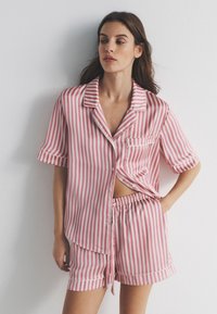 Vrouw draagt een roze-wit gestreept kort mouw knoophemd en bijpassende shorts, staand tegen een witte muur.