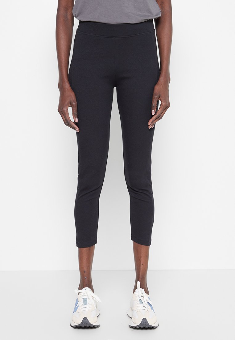 Leggings cortos negros fabricados con material elástico, con una cinturilla alta y combinados con zapatillas deportivas blancas. Textura suave, sin patrones visibles.