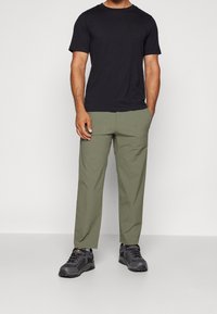 Sort t-shirt, rund hals; lys grønne bukser, afslappet pasform; grå vandresko med detaljer; stående figur med hænderne i lommerne.