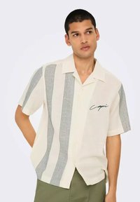 Camisa de manga corta color beige con rayas verticales negras y un estampado gráfico "Capri" en el lado izquierdo. Tejido ligero, corte relajado.