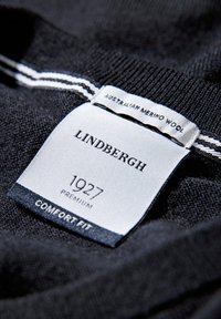 Czarna etykieta dzianinowa z napisem "LINDGREN", "1927" i "COMFORT FIT"; zawiera "AUSTRALIAN MERINO WOOL" w białym kolorze na tle z tkaniny.