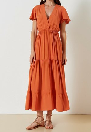 Kvinde iført en ærmeløs, dyb V-hals, lagdelt orange midi-kjole med flæseærmer og flade beige sandaler med ankelrem.