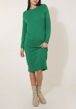Robe-sweater en tricot vert avec des manches longues, des poignets et un ourlet côtelés, présentant une coupe décontractée et une texture lisse. Associée à des bottines beiges ankle.