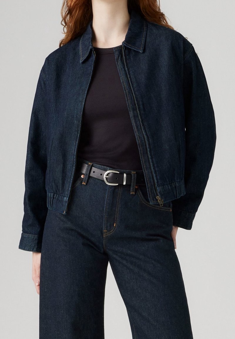 Femme portant une veste en jean foncé et un jean avec une ceinture noire et un haut côtelé noir, debout devant un fond clair uni.