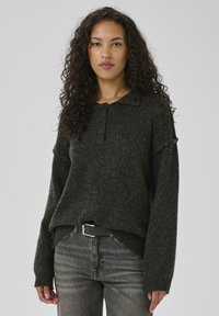 Woman with curly hair wearing a dark gray buttoned knit sweater and faded black jeans with a black belt, standing against a plain background.