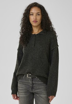 Woman with curly hair wearing a dark gray buttoned knit sweater and faded black jeans with a black belt, standing against a plain background.