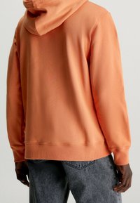 Orange hooded sweatshirt with a smooth texture, ribbed cuffs, and hem in a relaxed fit, paired with gray denim pants.